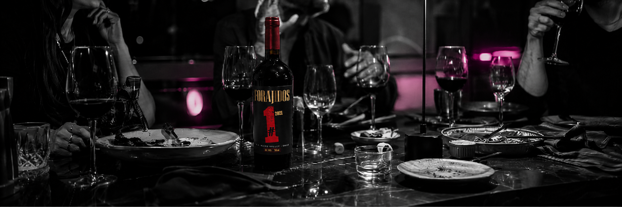 Mesa nocturna compartida con copas, platos y una botella Forajidos
